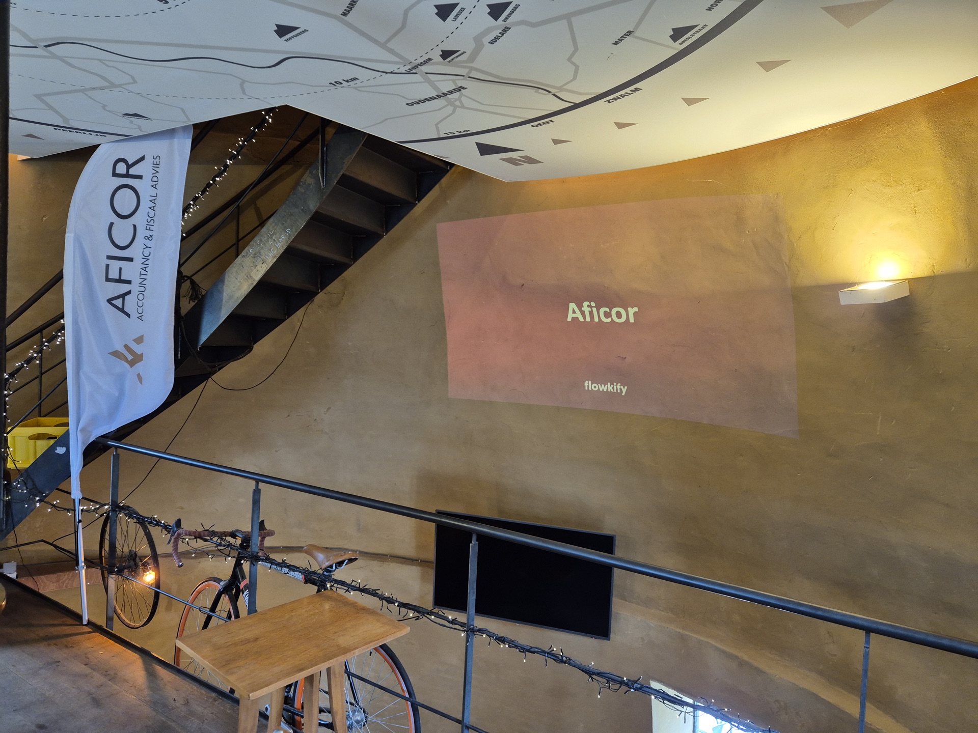 Aficor-banner naast een geprojecteerde slide in een zaal met leemwanden.