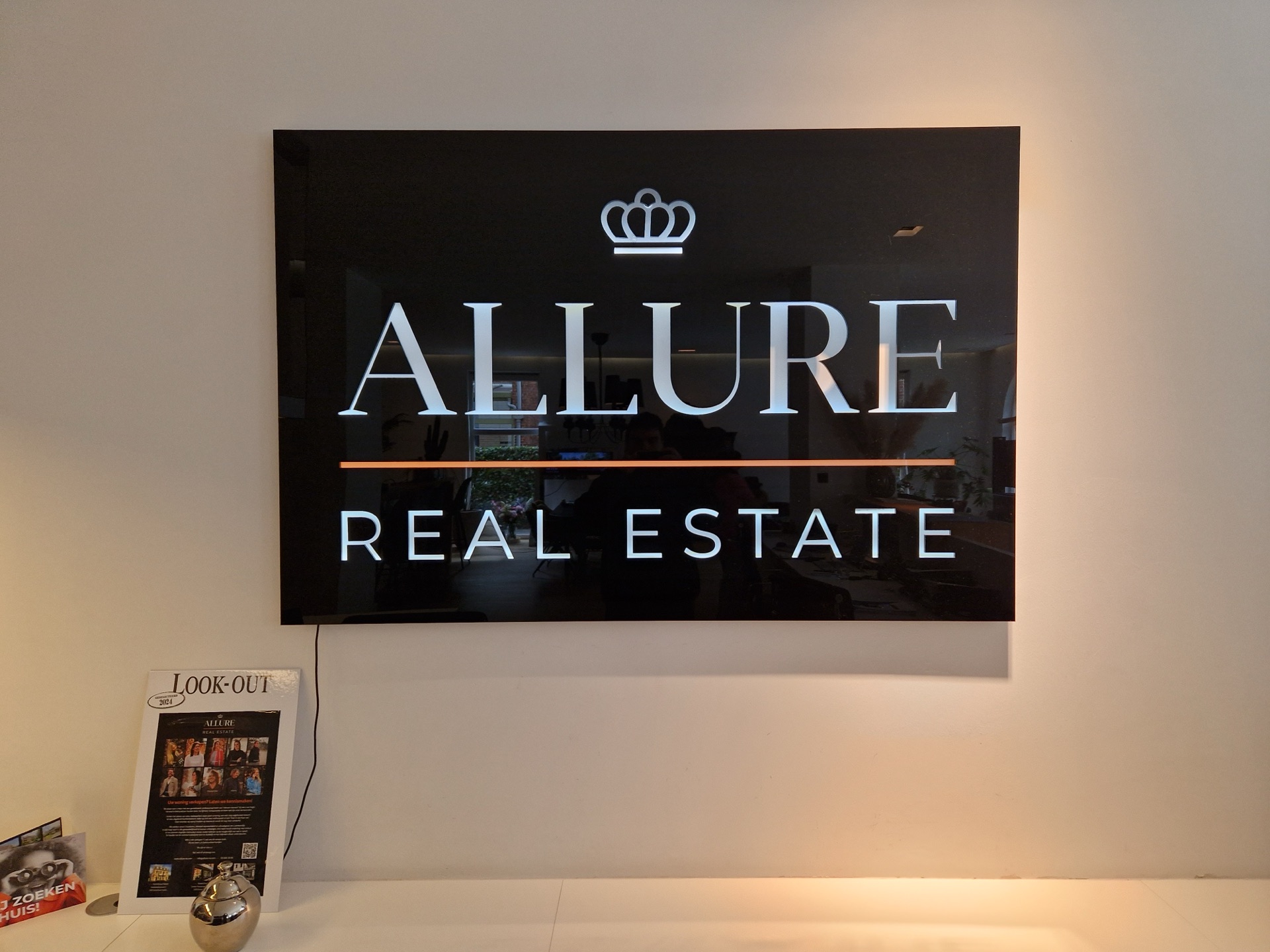 Verlicht bord met het logo van Allure Real Estate aan een kantoorwand.