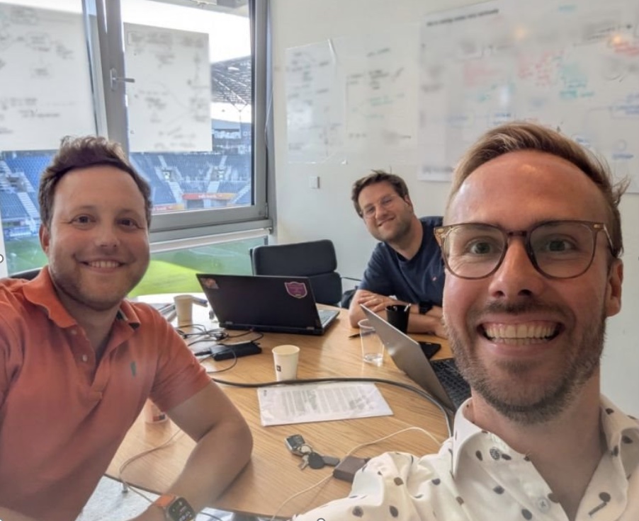 Selfie van drie mensen in een vergaderzaal met uitzicht op een stadion.