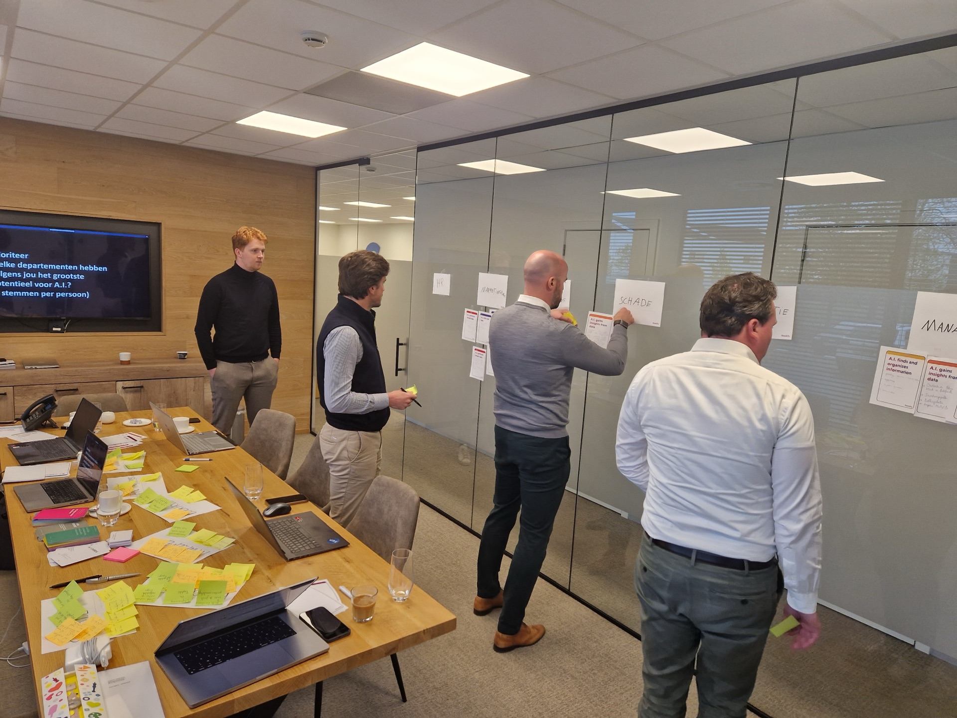 Deelnemers plakken papieren op een glazen wand, met post-its op de tafel.
