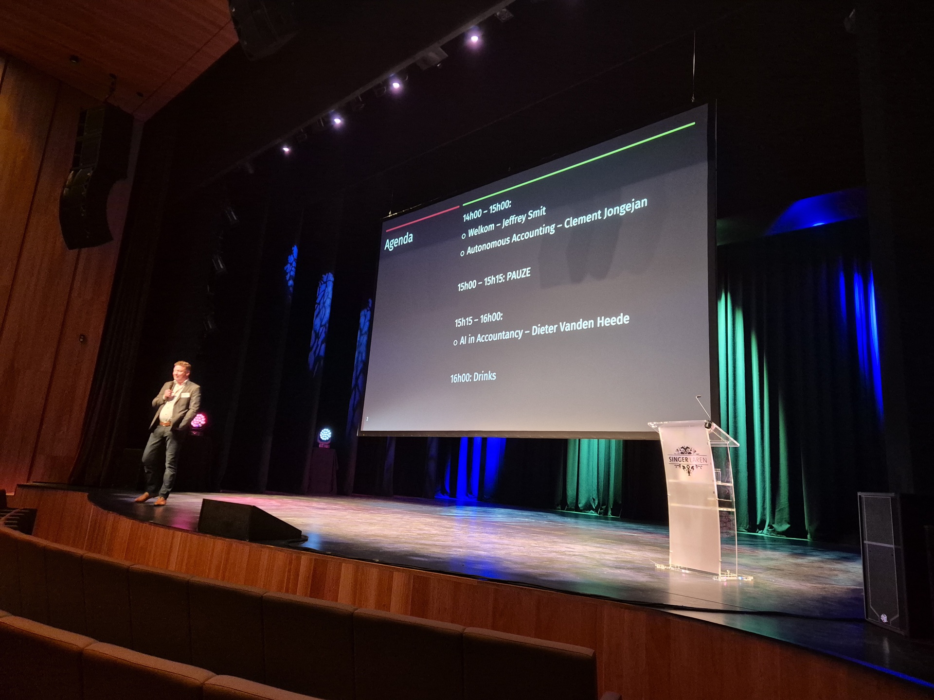 Spreker op een podium naast een groot scherm met de agenda van het symposium.